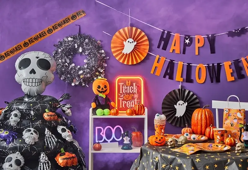 A Halloween-themed scene with carved pumpkins featuring glowing faces, lit candles on a table, and a person wearing a black outfit with a skeleton design alongside another person in a black cape. The background includes more pumpkins and festive decorations.